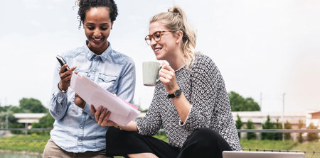 Zwei Frauen sitzen draußen, schauen auf einem Stapel Unterlagen und unterhalten sich.