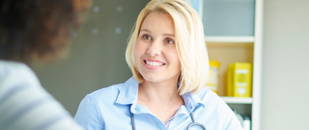 Eine Ärztin, die ein Stethoskop um den Hals hängen hat. Sie lächelt, sitzt einer weiteren Frau gegenüber und unterhält sich mit ihr.