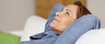 Lächelnde Frau, die ihre Arme hinter ihrem Kopf verkreuzt und ihren Kopf auf der Sofa-Lehne ablegt.